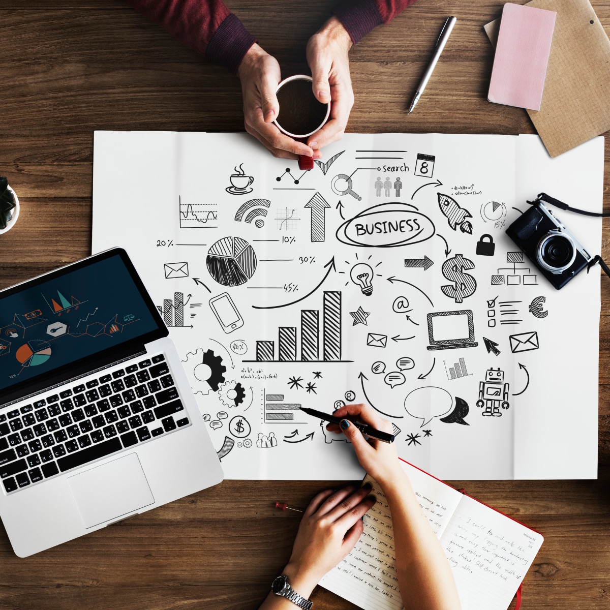 Two people brainstorming digital marketing strategies at a wooden desk with a sketch-filled paper showing graphs, charts, icons, and a laptop displaying analytics.