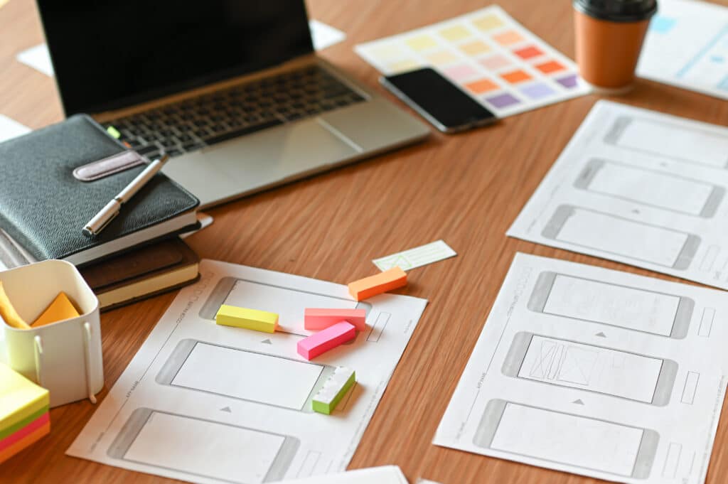 A wooden desk with wireframe sketches, sticky notes, a laptop, and design tools arranged for website planning and user interface development.
