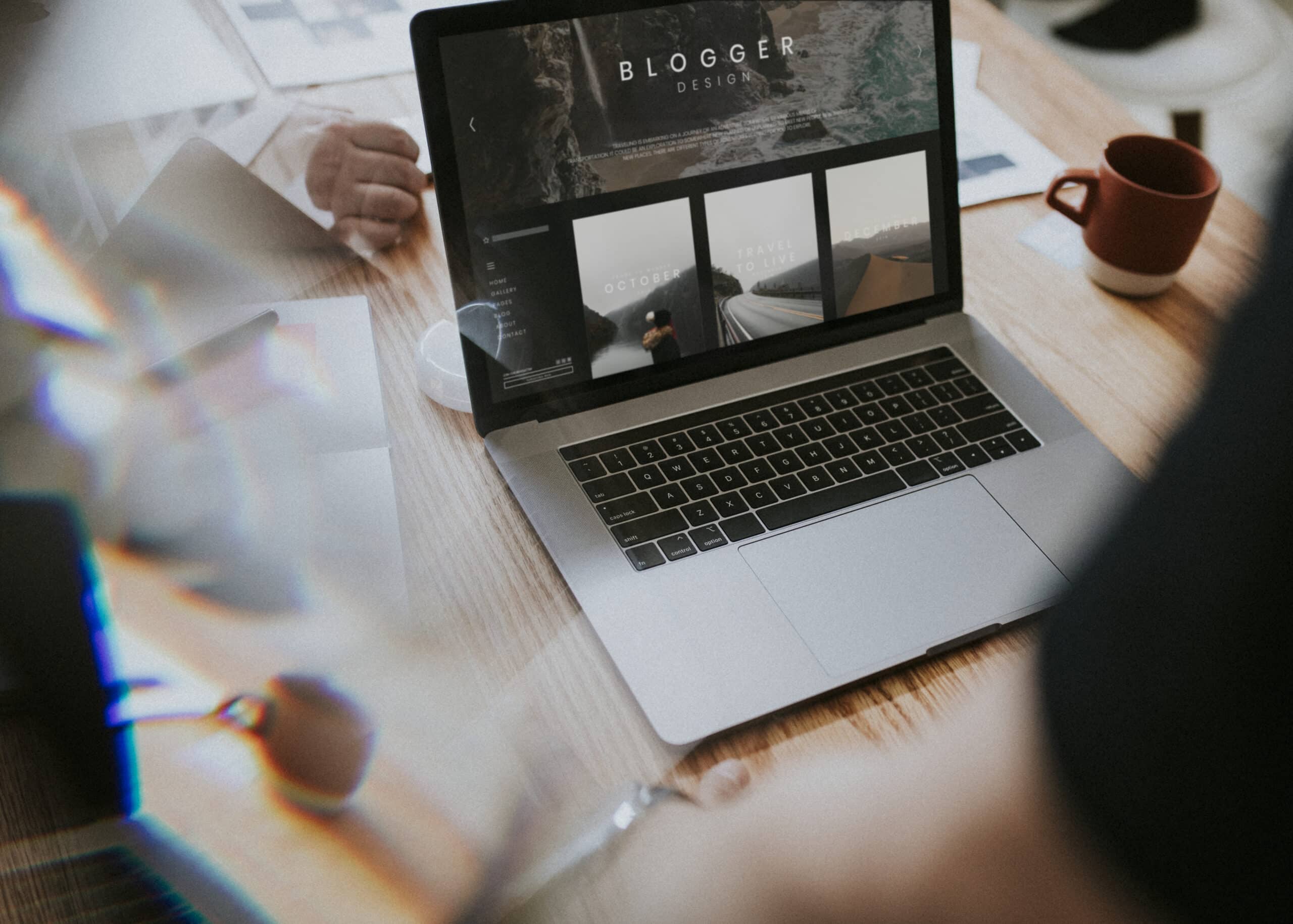 A laptop displaying a modern website layout sits on a wooden desk, surrounded by coffee mugs and blurred reflections, representing creative digital marketing workspace planning.