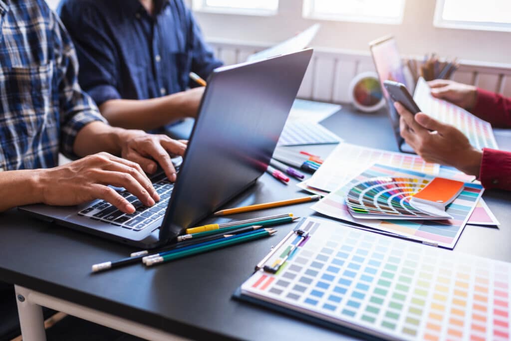 A group of designers working with laptops, smartphones, and colour swatches at a desk, collaborating on a creative graphic design project with vibrant palette selections and tools.