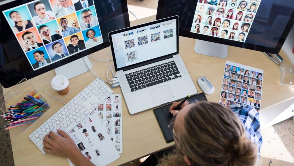 A website design working at a multi-monitor setup with a laptop, graphic tablet, and image assets, editing and organising profile photos for a website interface or portfolio layout.