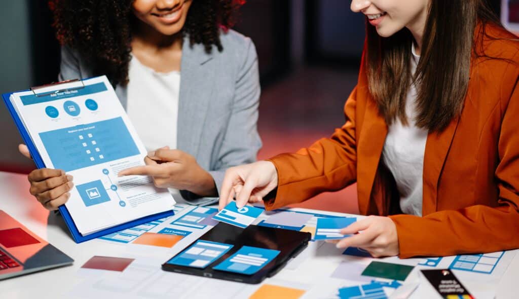 Two female designers brainstorming over a web design layout using paper mockups and wireframes on a table, planning user interface and structure for a digital project.